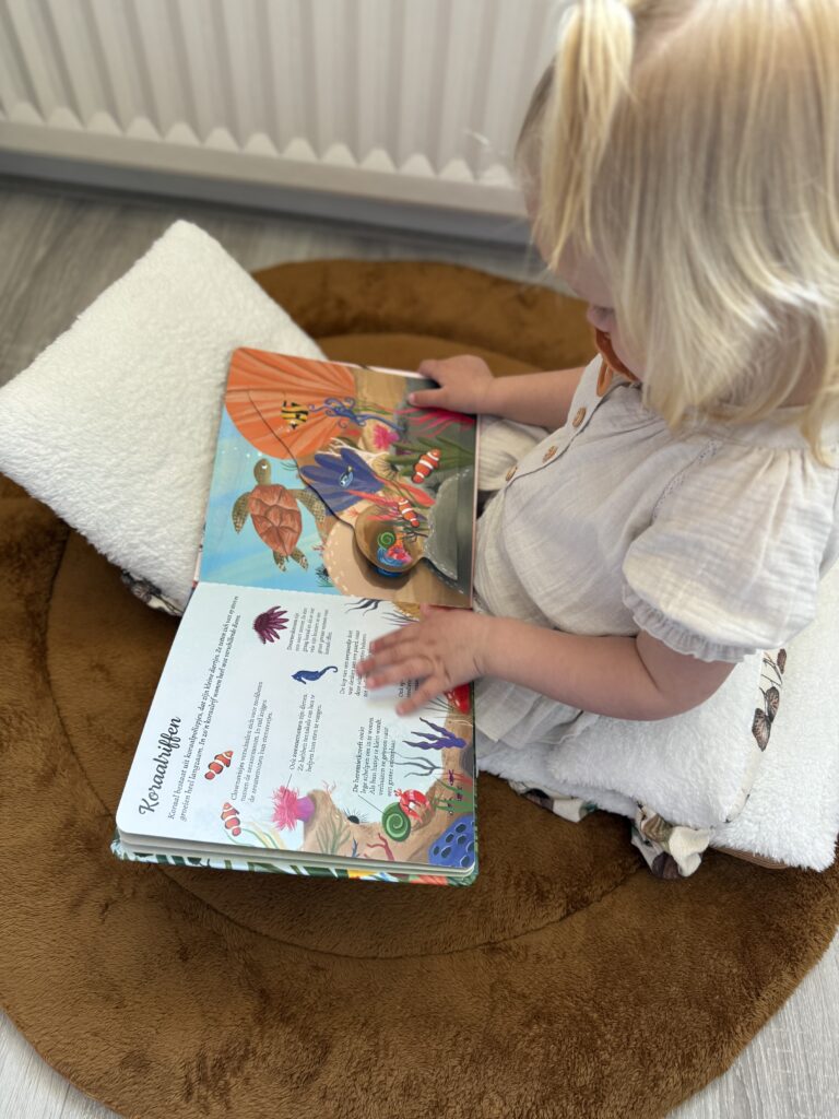Peuter zit op een zacht kussen en bladert geconcentreerd door het kleurrijke flapjesboek Ik ontdek de natuur.