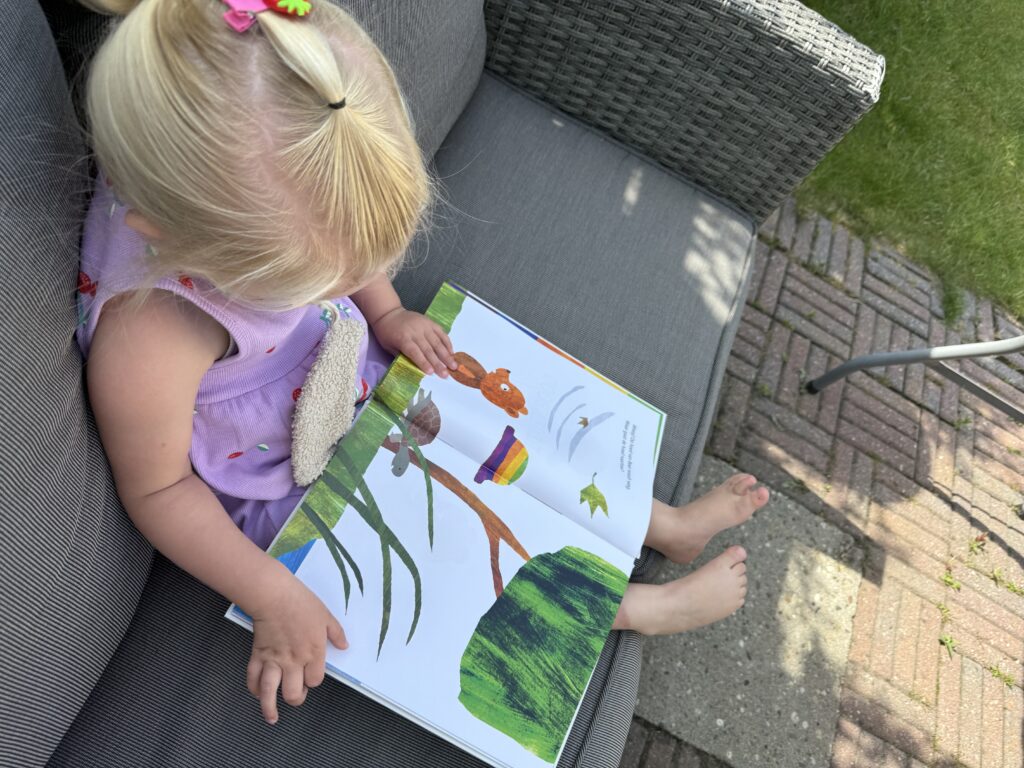 Peuter leest aandachtig het kleurrijke boek Regenboog hoedje tijdens een zonnig moment op de tuinbank.