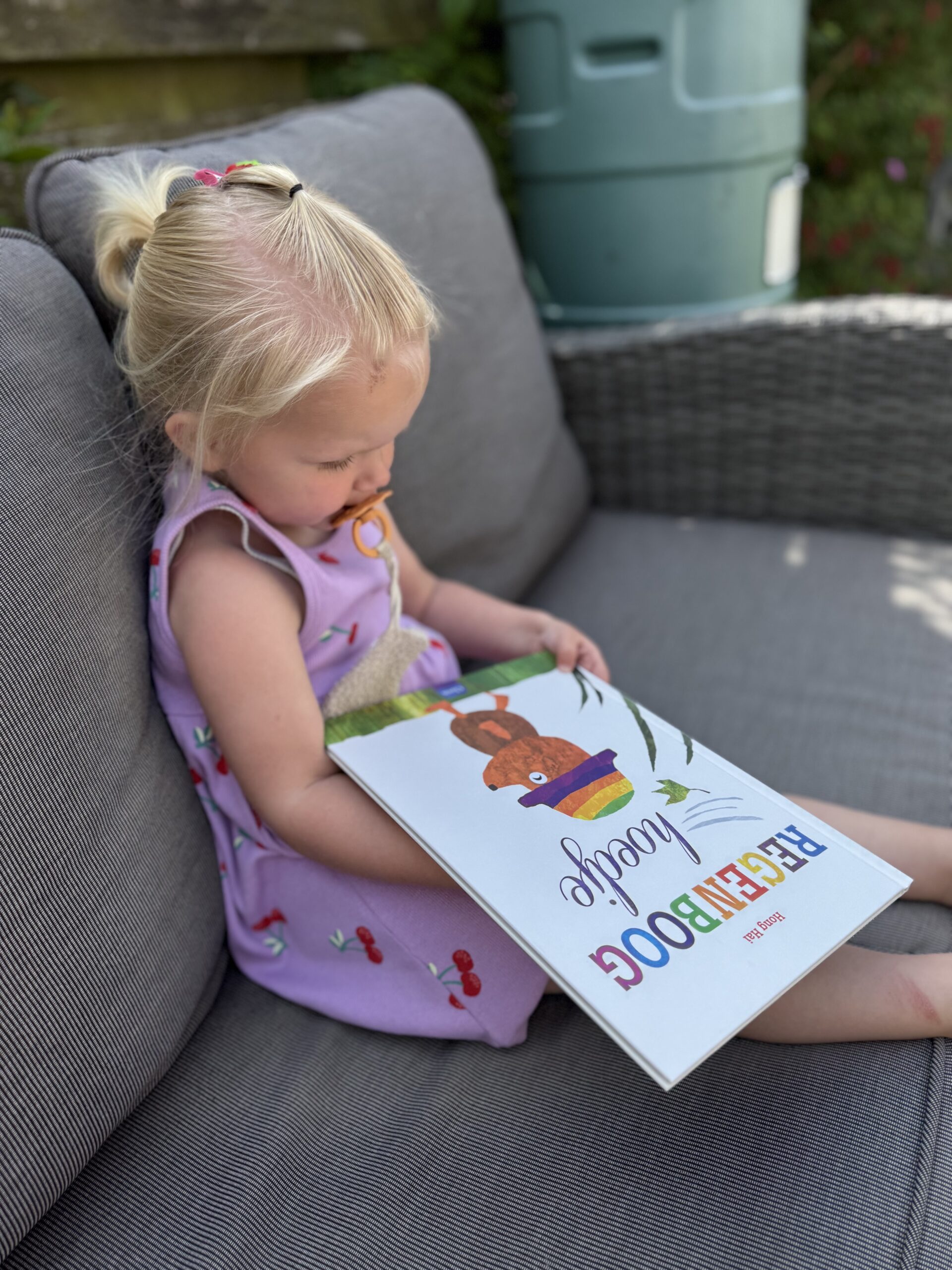 Kleuter bekijkt de cover van Regenboog hoedje vol nieuwsgierigheid tijdens een rustig leesmoment buiten.