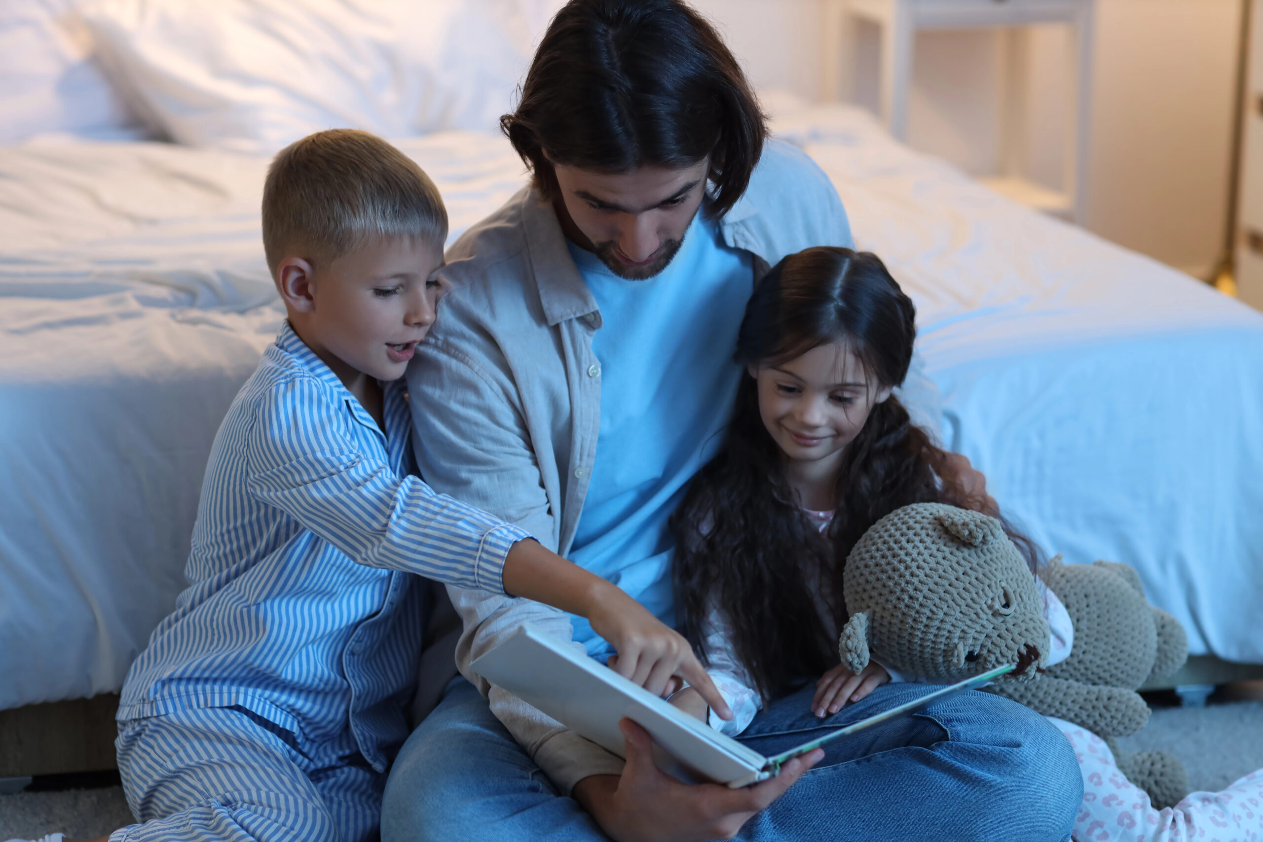 Vader leest voor aan zijn twee kinderen in pyjama, samen knus op de slaapkamer met een boek en een knuffel.